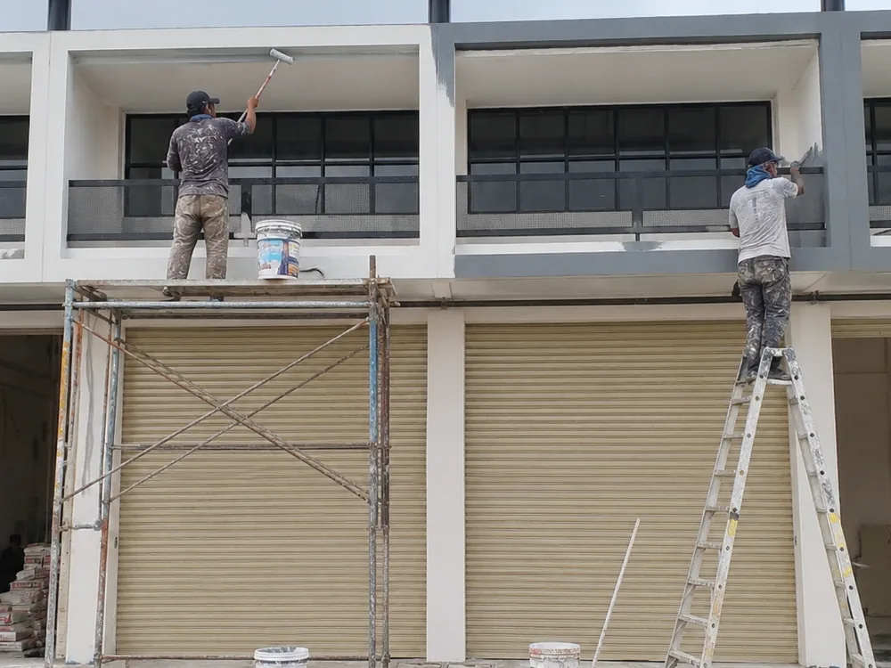 Dua tukang ASATURA mengecat fasad rumah dua lantai dari scaffolding